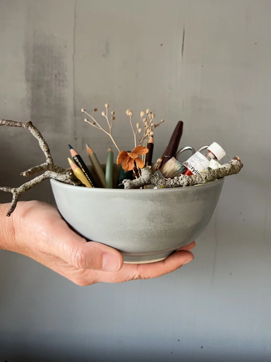 Eine Hand hält eine Keramikschale mit Farbstiften, Pinsel, Farbtube, Zweigen und getrockneten Blüten vor einem ruhigen Hintergrund